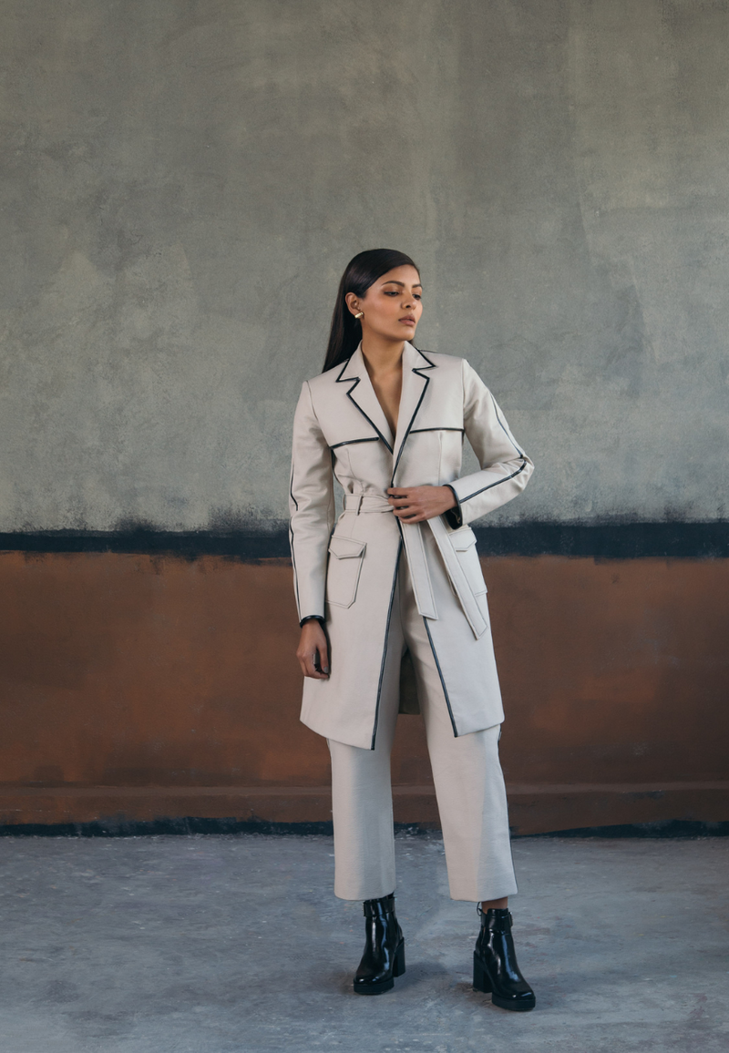 This back-pleated leather trench jacket in beige is crafted from supple rexine and lined with luxe poly-satin. This knee-length trench coat is detailed with pleating on the back, black leather piping, cargo pockets and a leather belt for fastening. Style it with a pair of pants or jeans along with boots/booties in fall and winter.