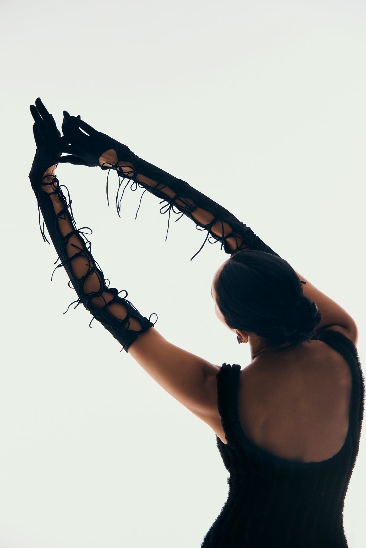A person viewed from behind wearing long black lace-up gloves with fringe, posed with arms raised against a plain light background.